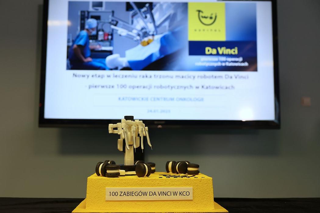 Na pierwszym planie żółto-brązowy tort z etykietą „100 zabiegów Da Vinci w KCO”, w tle prezentacja o operacjach wykonywanych przez robota