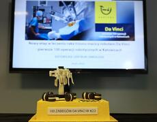 Na pierwszym planie żółto-brązowy tort z etykietą „100 zabiegów Da Vinci w KCO”, w tle prezentacja o operacjach wykonywanych przez robota